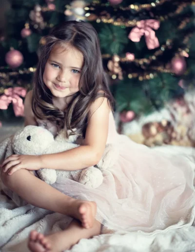 Young girl in a pink dress holding a teddy bear beside a Christmas tree
